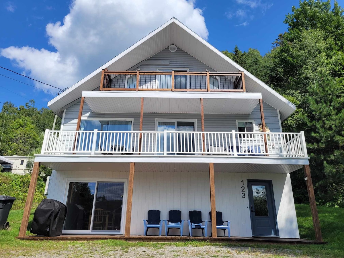 The three-story chalet features a covered balcony on the upper level, providing space for relaxation. The front entrance is highlighted by a porch area with several dark chairs arranged neatly. Large windows offer ample natural light, with a view of the surrounding greenery.