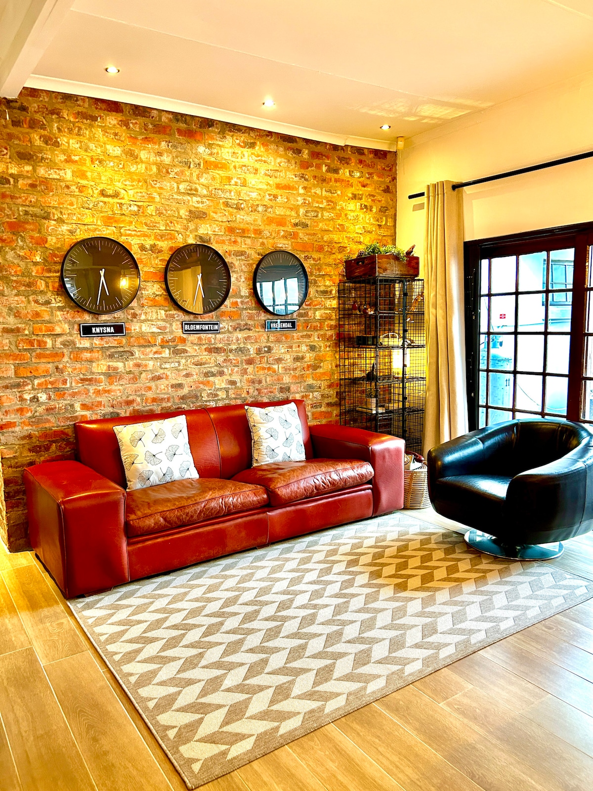A seating area features a red leather sofa adorned with two patterned pillows, complemented by a sleek black chair. The exposed brick wall adds texture, while three wall clocks indicate different times. A geometric rug anchors the space, and large windows allow natural light to illuminate the room.