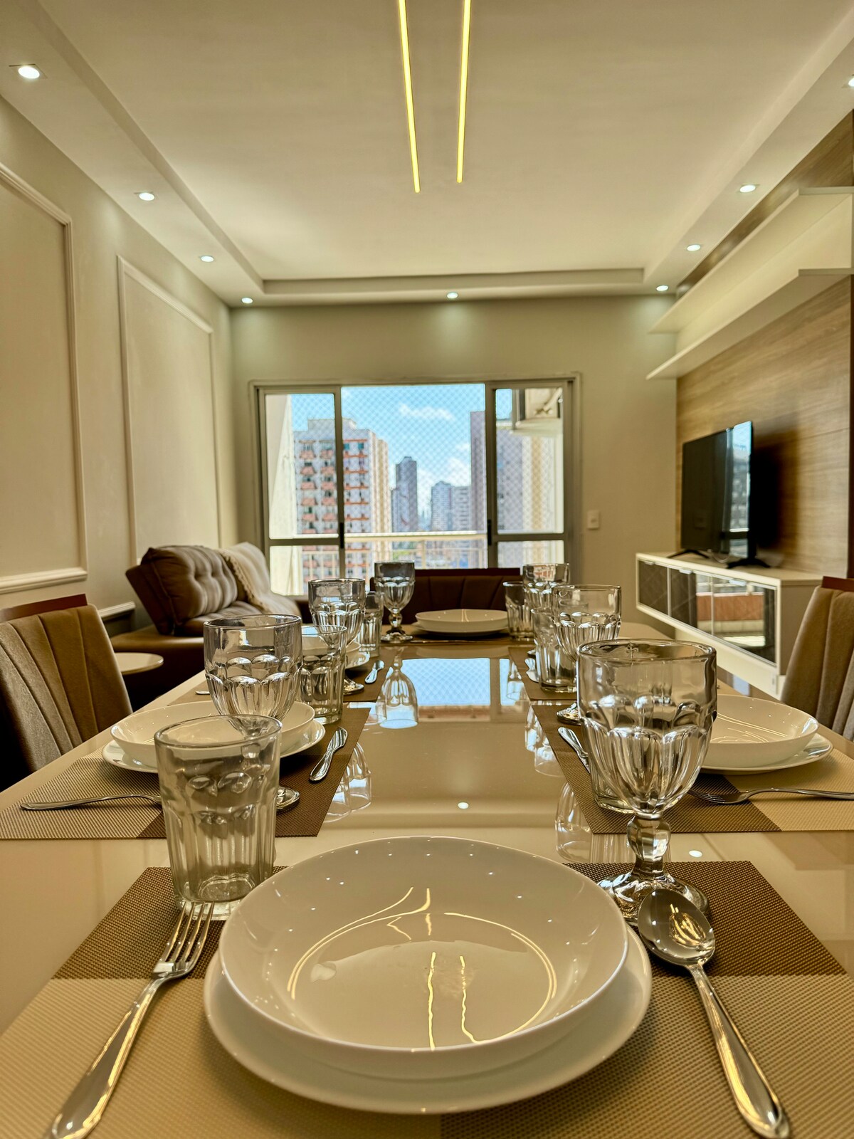 A modern dining area is set with a glass table, neatly arranged with dishes, glasses, and utensils. The view of the city skyline is visible through large windows in the background. The space is illuminated by soft lighting, creating an inviting atmosphere.