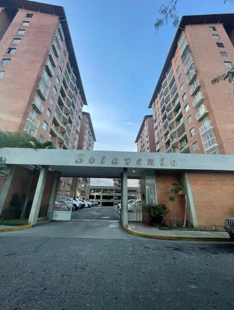 The entrance of the Sotavento complex is framed by two tall brick buildings. A wide driveway leads into a parking area, flanked by greenery and featuring a decorative sign that reads 'Sotavento.' Clear skies are visible above, contributing to the serene atmosphere.