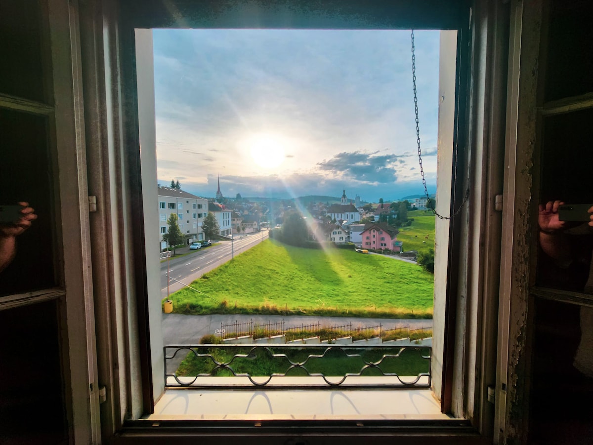 A view from the window showcases a serene landscape, featuring rolling green fields under a sunset sky. The distant buildings and mountains create a picturesque backdrop, while the gentle light enhances the peaceful atmosphere outside.