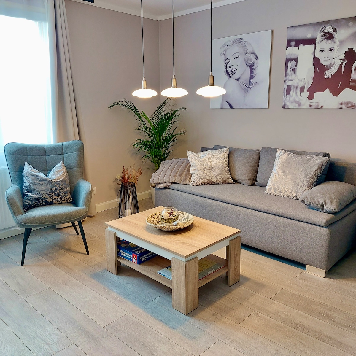 A welcoming living area is furnished with a gray sofa and light wooden coffee table. A potted plant adds a touch of greenery, while two pendant lights hang above. Decorative art pieces adorn the walls, complementing the neutral color scheme.