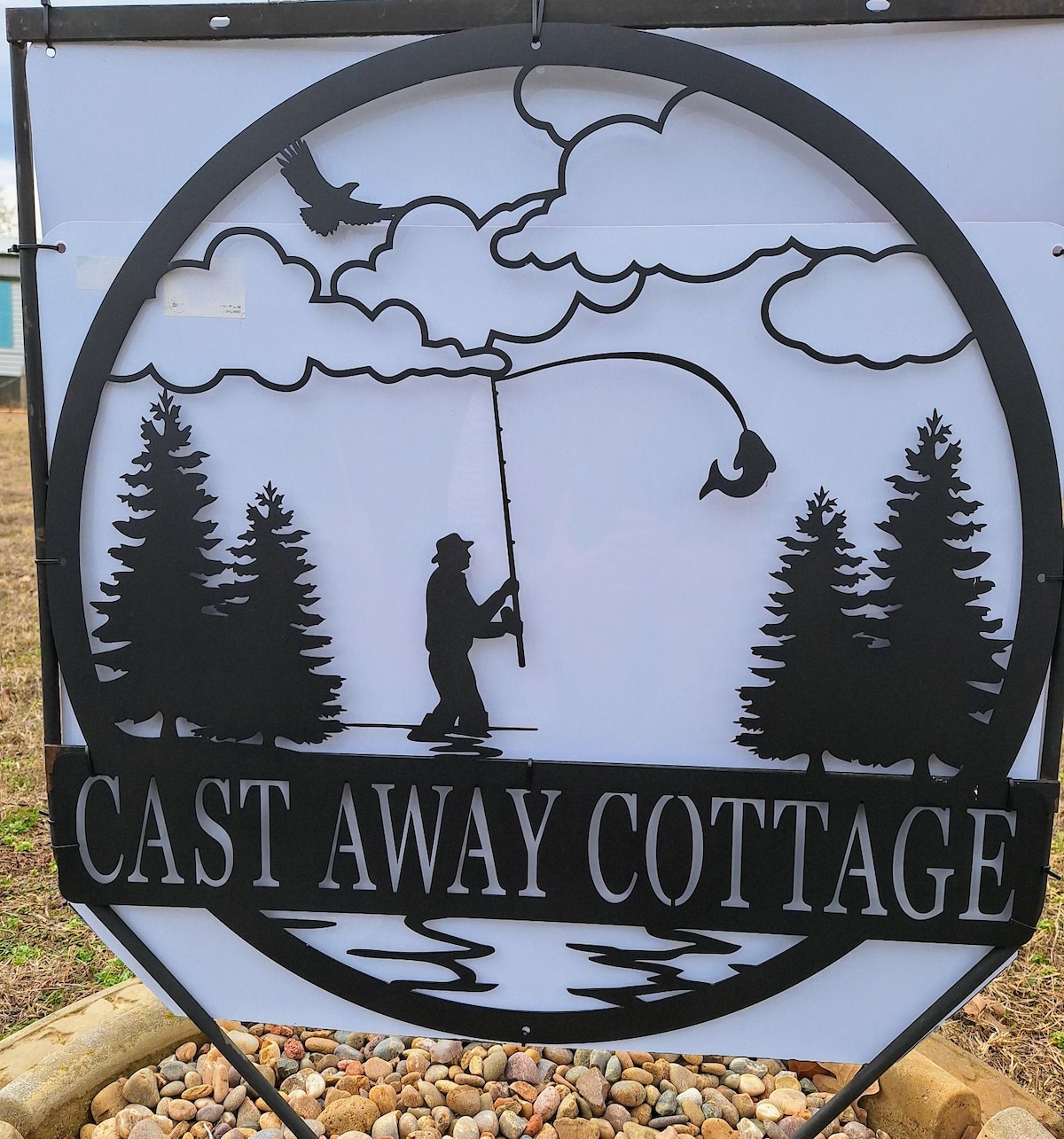 A decorative sign for Cast Away Cottage features a silhouette of a fisherman casting a line under a cloudy sky. Surrounding pine trees frame the scene, which is set above a stylized water line, creating a serene representation of lake life.