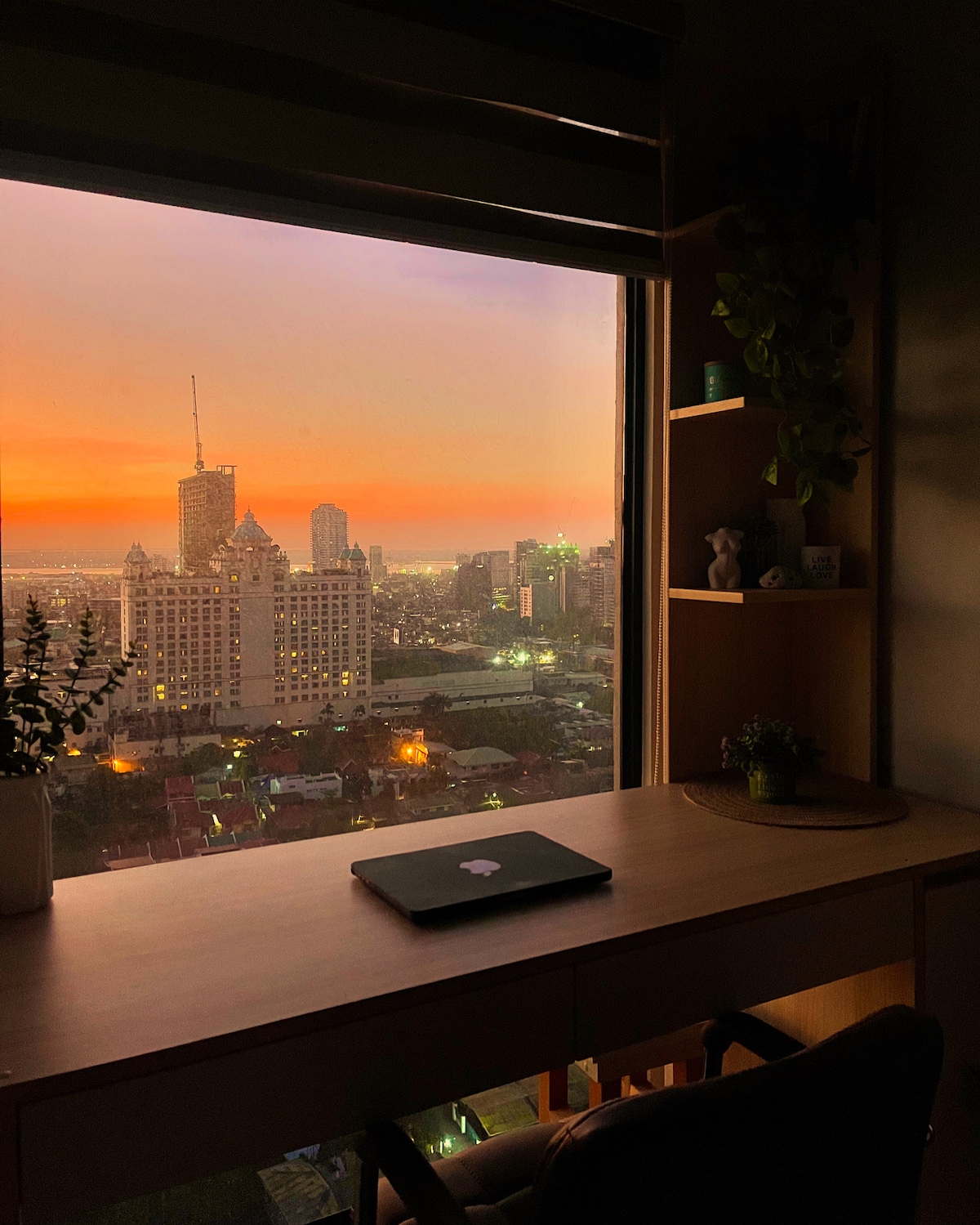A cozy workspace is positioned beside a large window, offering a stunning view of the city during sunset. Soft orange and pink hues illuminate the skyline, while a laptop sits on a sleek wooden desk adorned with small potted plants, enhancing the serene atmosphere.