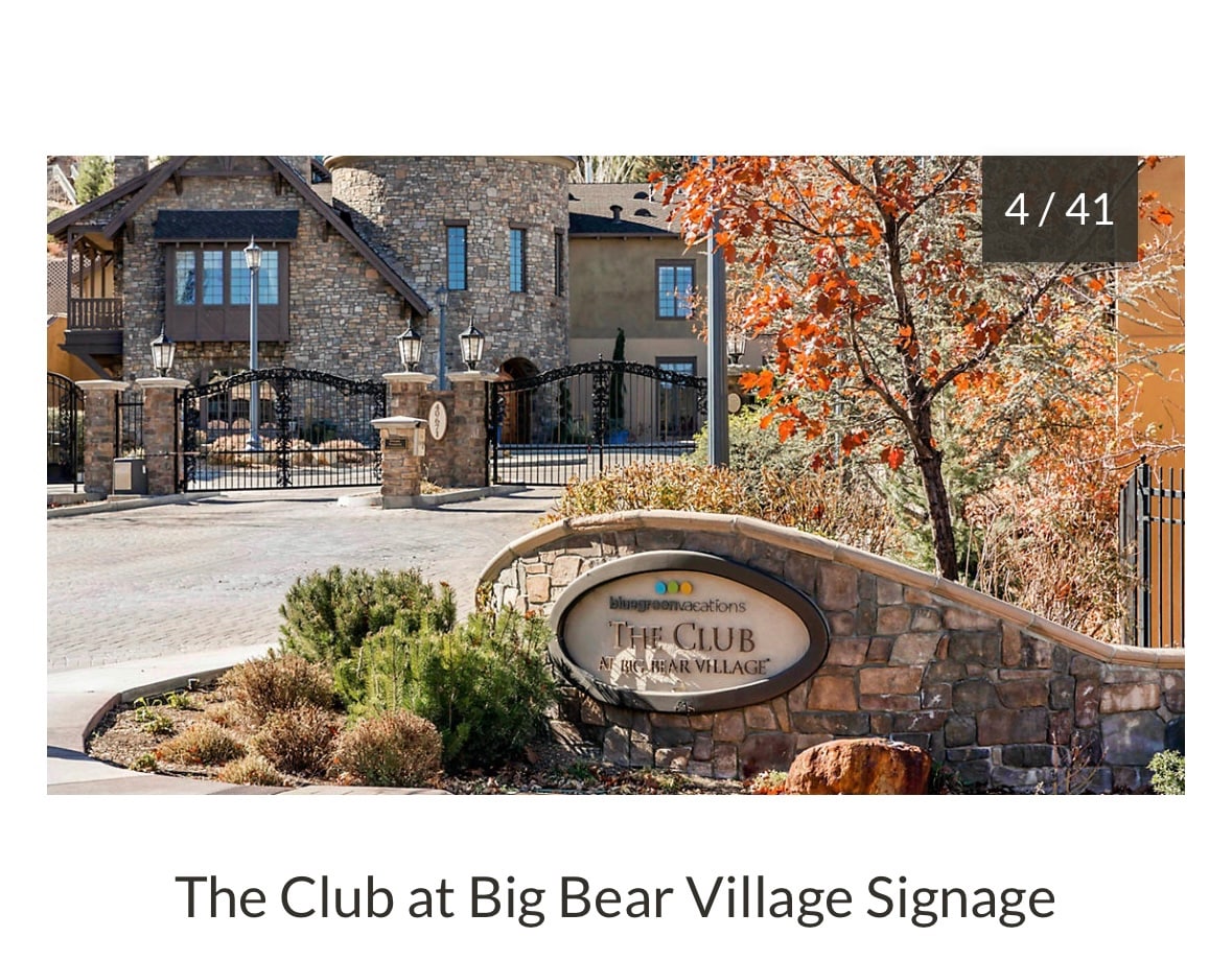 A stone structure features a welcoming entrance marked by a prominent sign displaying 'The Club at Big Bear Village.' Lush greenery and seasonal foliage surround the entryway, contributing to an inviting ambiance, while decorative lanterns frame the entrance path.