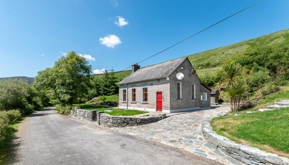 A charming stone exterior of a house is set against a backdrop of rolling green hills. A red door adds a pop of color, while a stone path leads to the entrance. Lush greenery surrounds the property, creating a sense of tranquility and connection to nature.