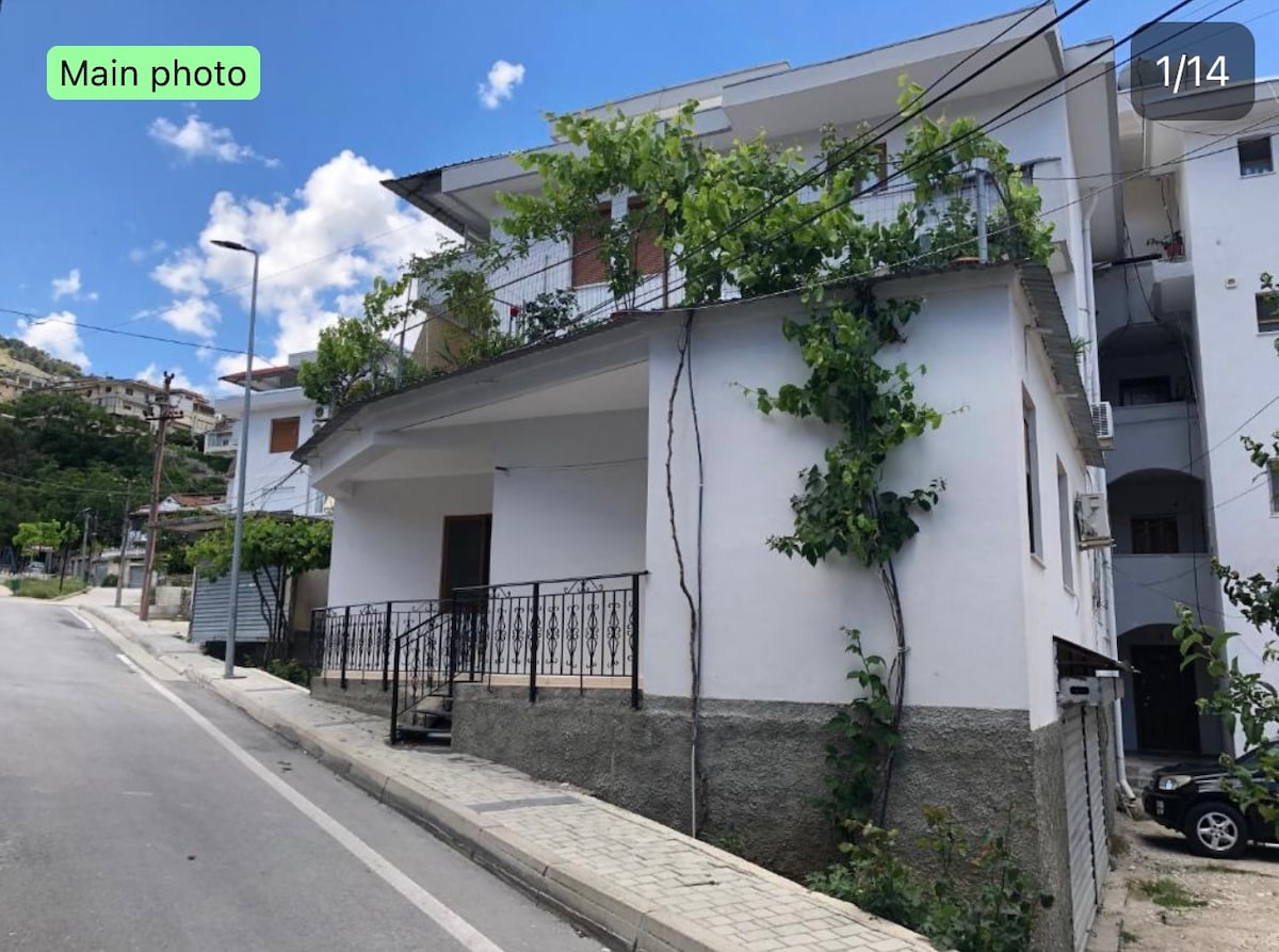 A two-story building features a white exterior with a balcony adorned with climbing greenery. A stone pathway leads to the entrance, while nearby accommodations are visible in the background. The area is well-lit by street lamps, and the sky above is partly cloudy.