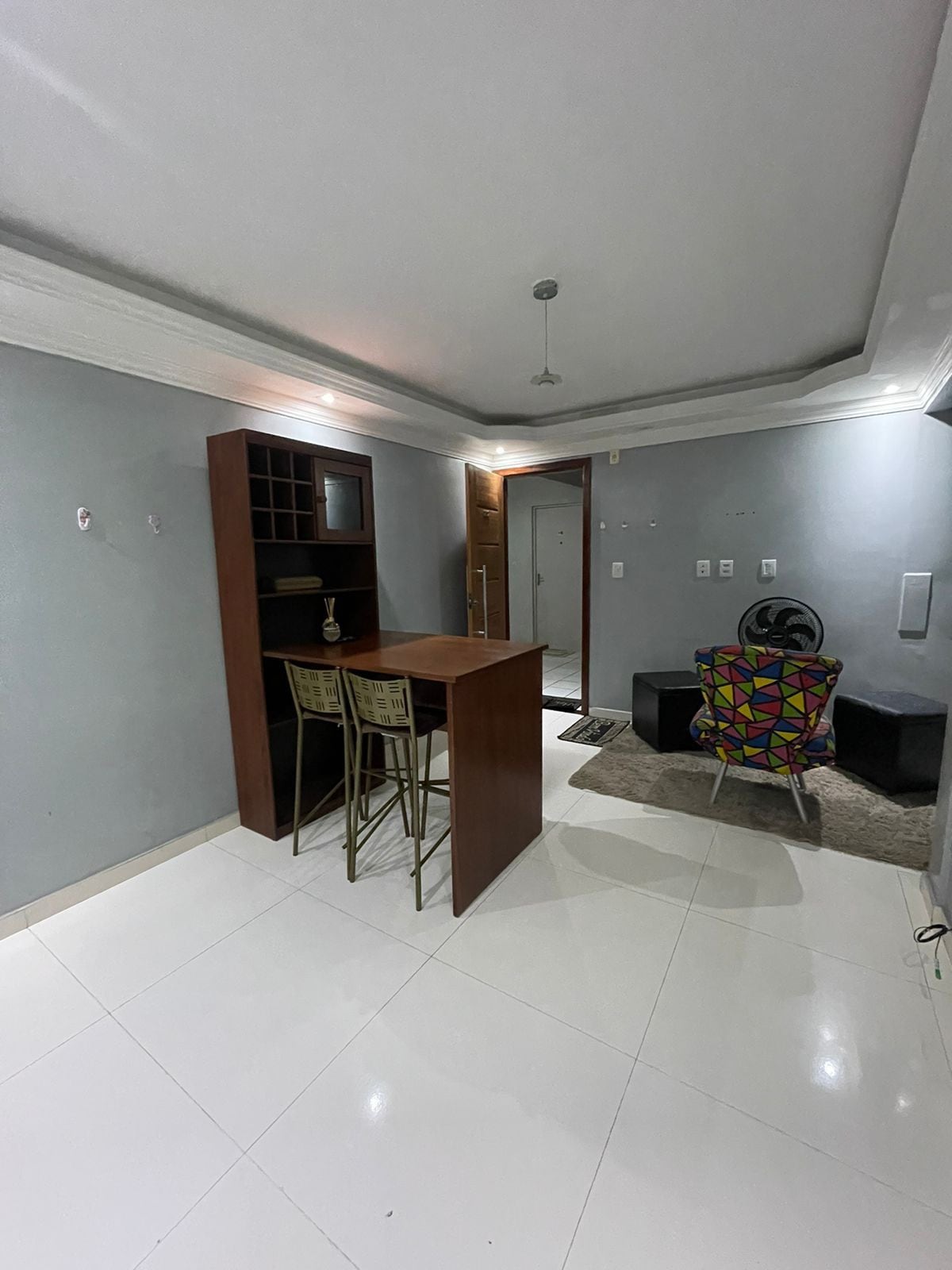 A well-lit living area features a modern bar table with two stools, positioned beside a colorful accent chair. The walls are a soft gray, and the light tile flooring enhances the spacious feel. A doorway leads to the entrance, while a small rug adds warmth.