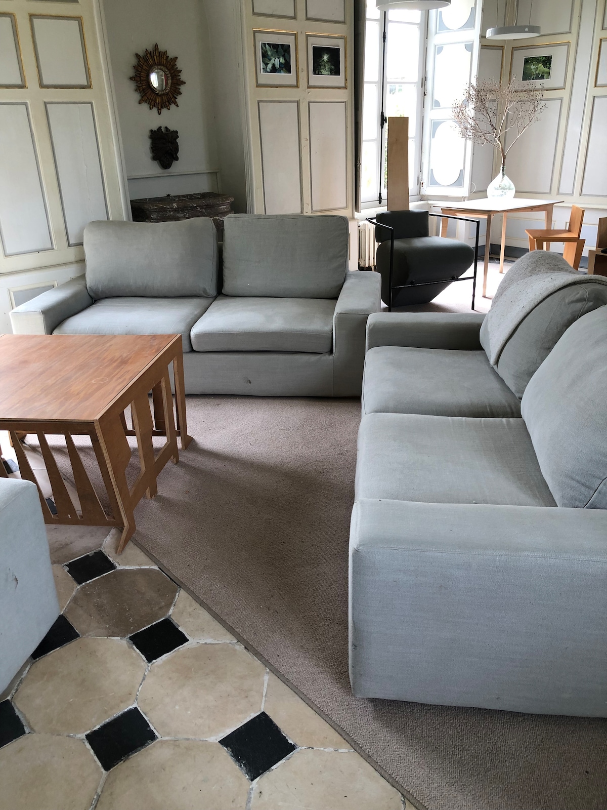A comfortable seating area features two light gray sofas arranged around a wooden coffee table. Natural light enters through large windows, illuminating the soft textures of the furniture and the patterned floor. Decorative elements and a dining table can be seen in the background.