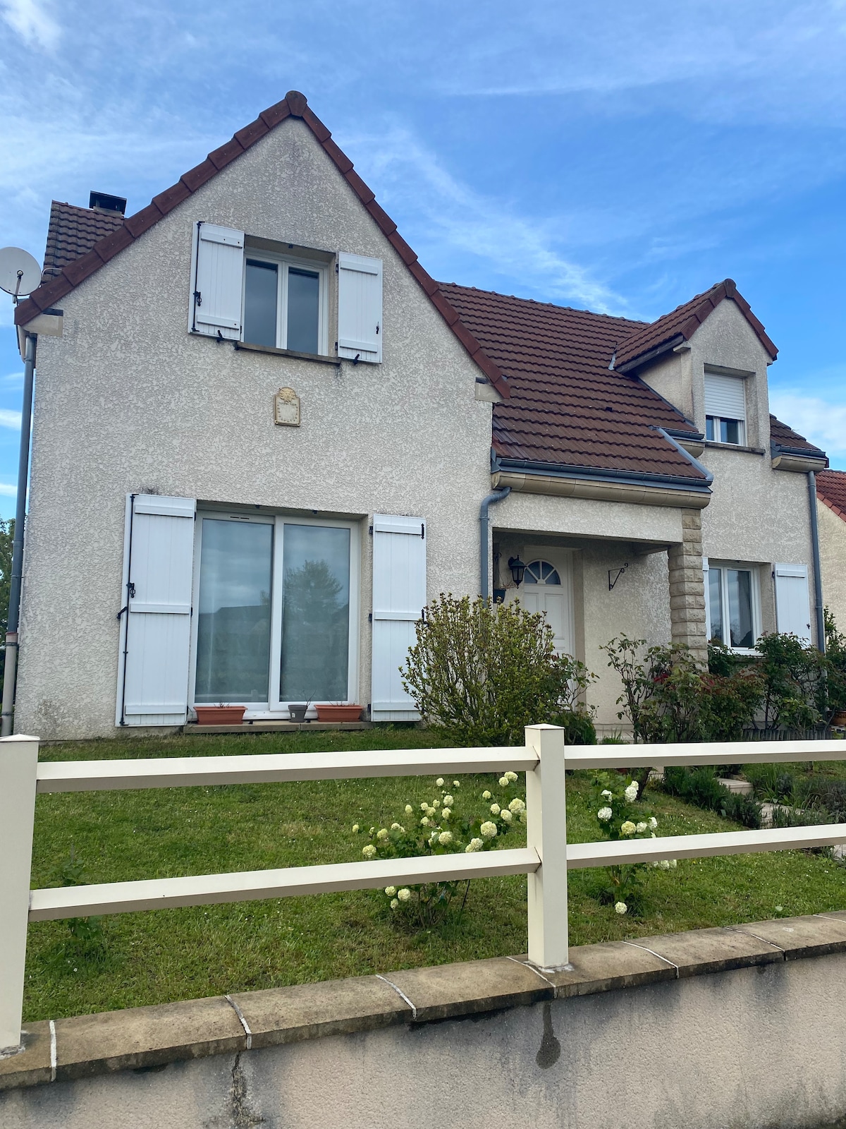 A light-coloured two-story house is depicted, featuring a sloped roof and white shutters. The facade is complemented by a small garden with green grass and flower beds. Large windows are visible, allowing natural light into the living spaces.