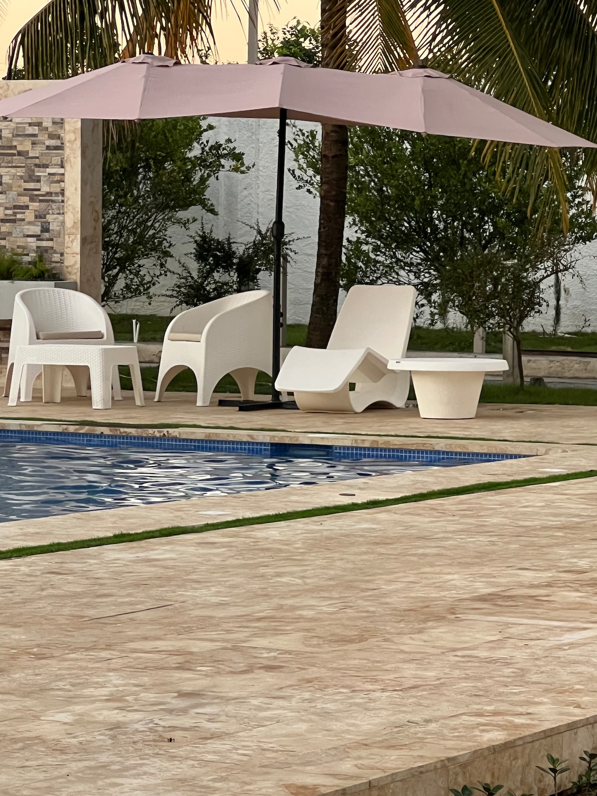 A serene outdoor pool area features a swimming pool bordered by light-colored tiles. Two white lounge chairs and a small table are positioned under a large pink umbrella, providing shade and a relaxing space surrounded by lush greenery.