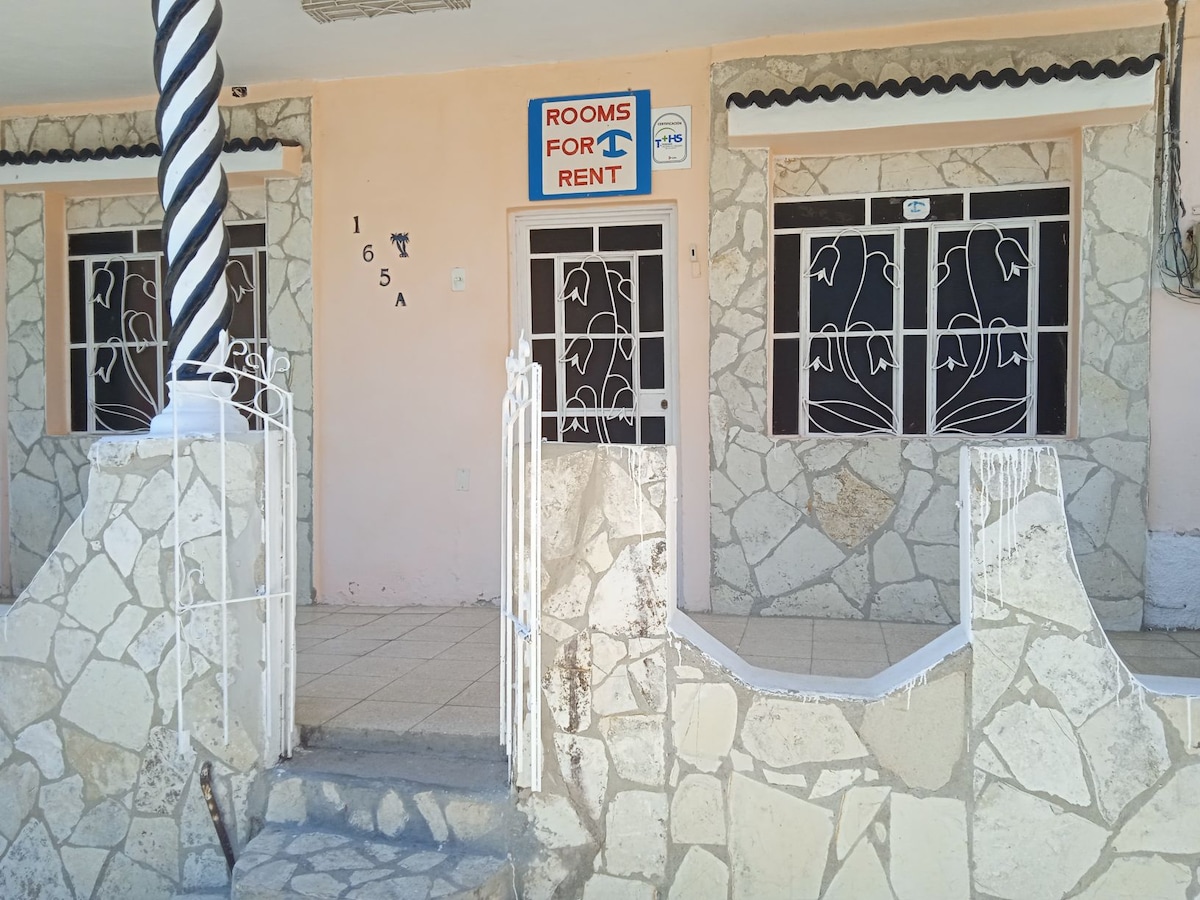 The exterior of a rental property is shown, featuring a stone facade and a decorative fence. Large windows adorned with floral ironwork allow natural light into the rooms. A sign reading 'Rooms for Rent' is prominently displayed above the entrance.