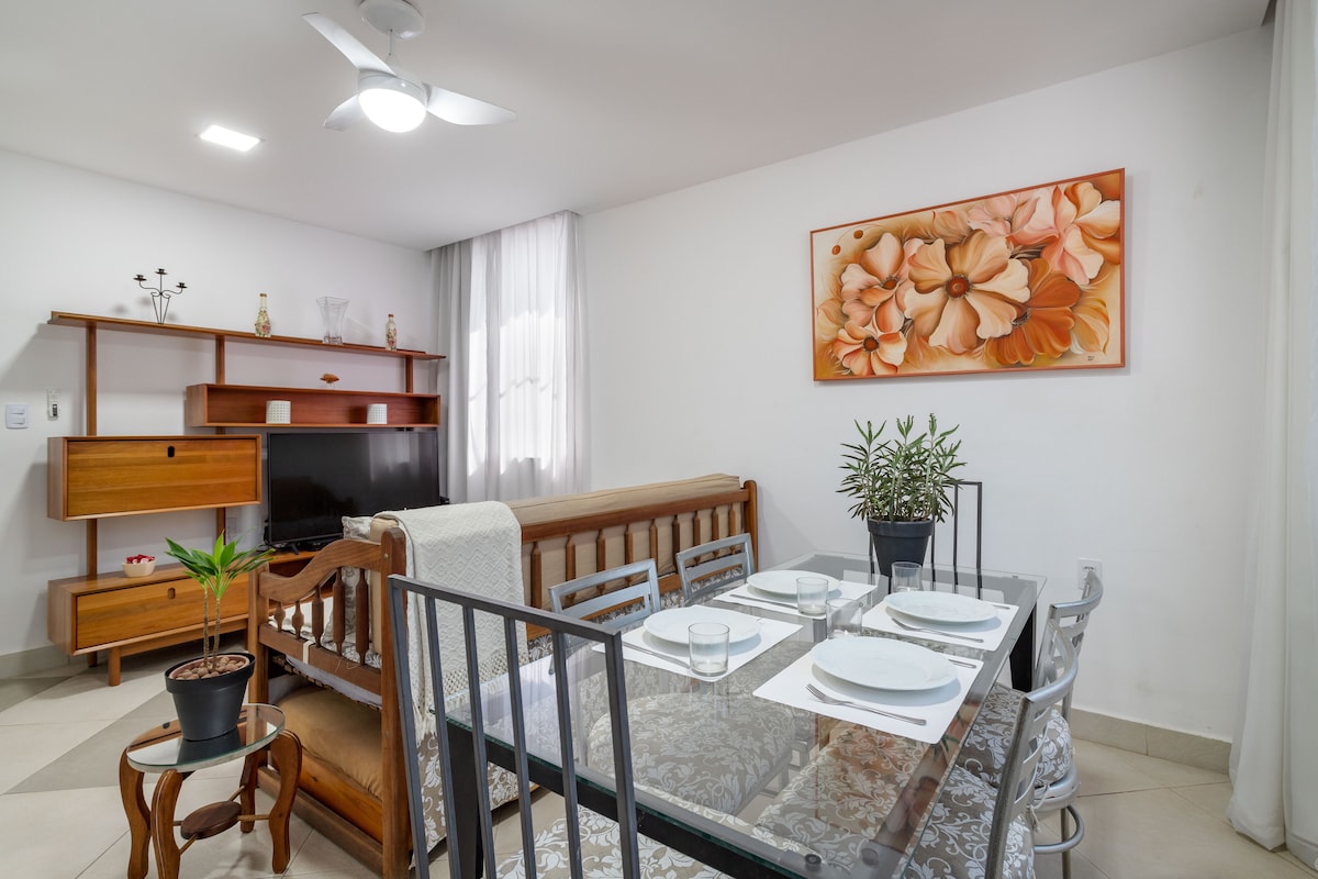 The living area is furnished with a wooden sofa and a dining table set for four, featuring elegant tableware. A wall-mounted television is positioned on a wooden shelf, and a floral artwork adds vibrancy to the space. Natural light enters through sheer curtains.
