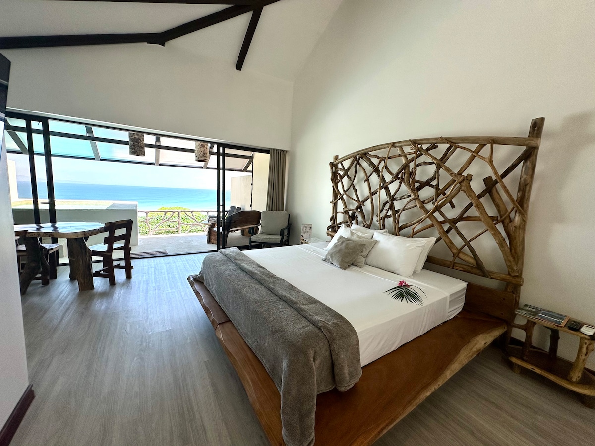 A spacious room features a large wooden bed with a textured headboard. Natural light floods in from expansive glass doors, revealing a view of the ocean. A dining table with chairs is positioned nearby, extending the inviting indoor space.