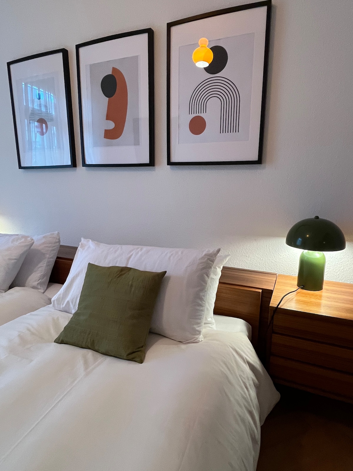A well-appointed sleeping area features two spacious beds with crisp white linens and decorative pillows. Beside each bed stands a wooden nightstand with a modern green lamp, which provides a warm glow. Three framed art pieces are displayed above the beds, adding a touch of character.