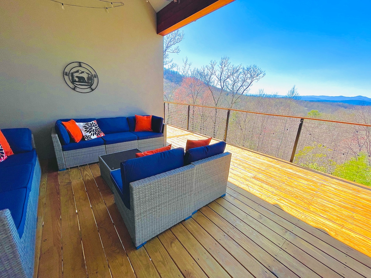 A spacious outdoor deck features comfortable seating arrangements with blue cushions and orange accents. The wooden deck opens up to panoramic views of the surrounding mountains and treetops under a clear blue sky.