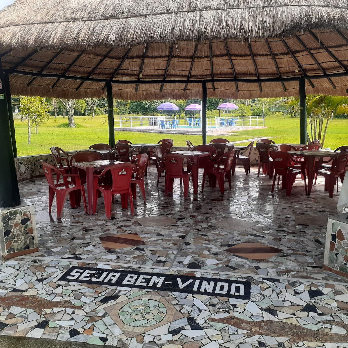 An outdoor seating area features a thatched roof, surrounded by lush greenery. Red plastic chairs are arranged around tables on a decorative stone floor. The words 'SEJA BEM-VINDO' are displayed prominently at the center. A glimpse of the pool area can be seen in the background.