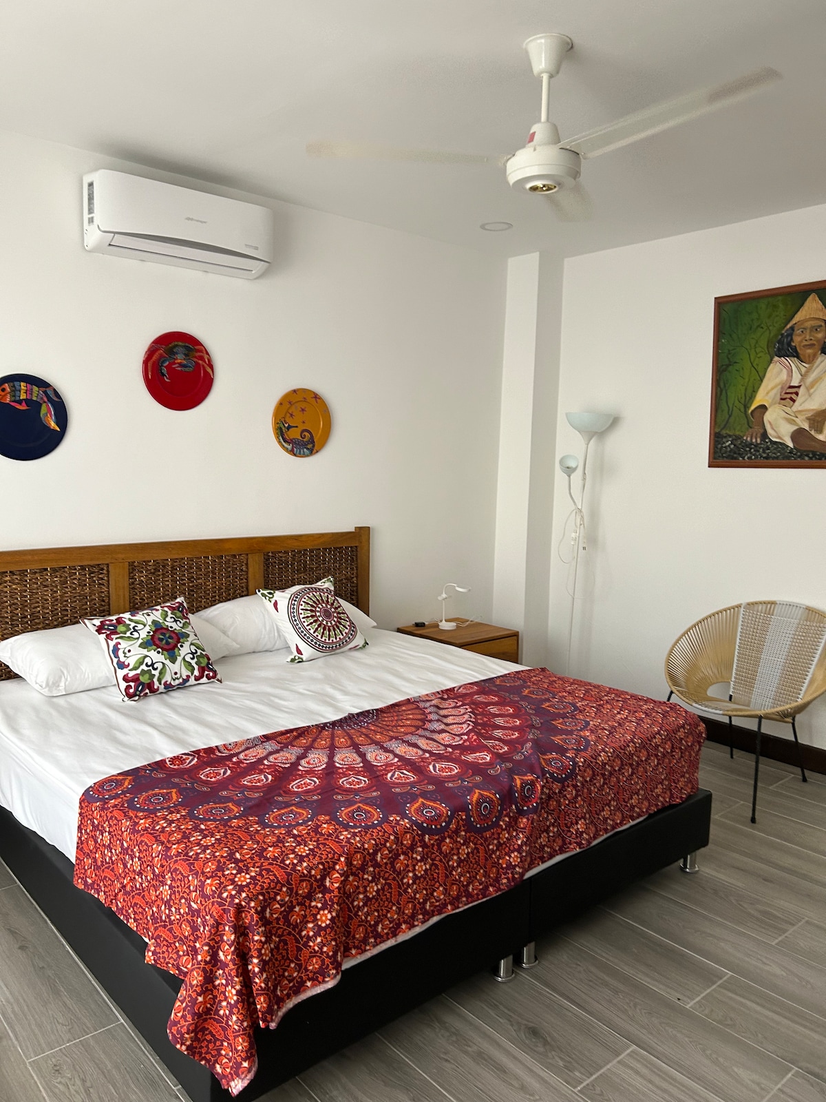 The sleeping area features a spacious bed with a colorful patterned blanket. Wall decorations add a touch of character, while a fan and air conditioning unit are visible for comfort. Natural light fills the room through a nearby window, enhancing the clean, modern aesthetics.