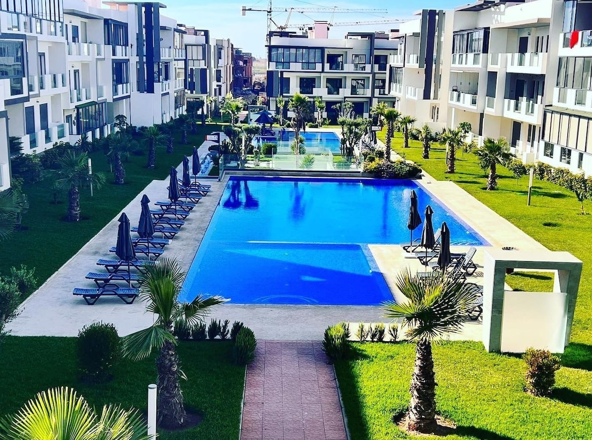 A vast outdoor area features a large swimming pool surrounded by neatly arranged sun loungers. Lush greenery and palm trees enhance the serene environment, while modern apartment buildings frame the scene, creating a peaceful retreat.