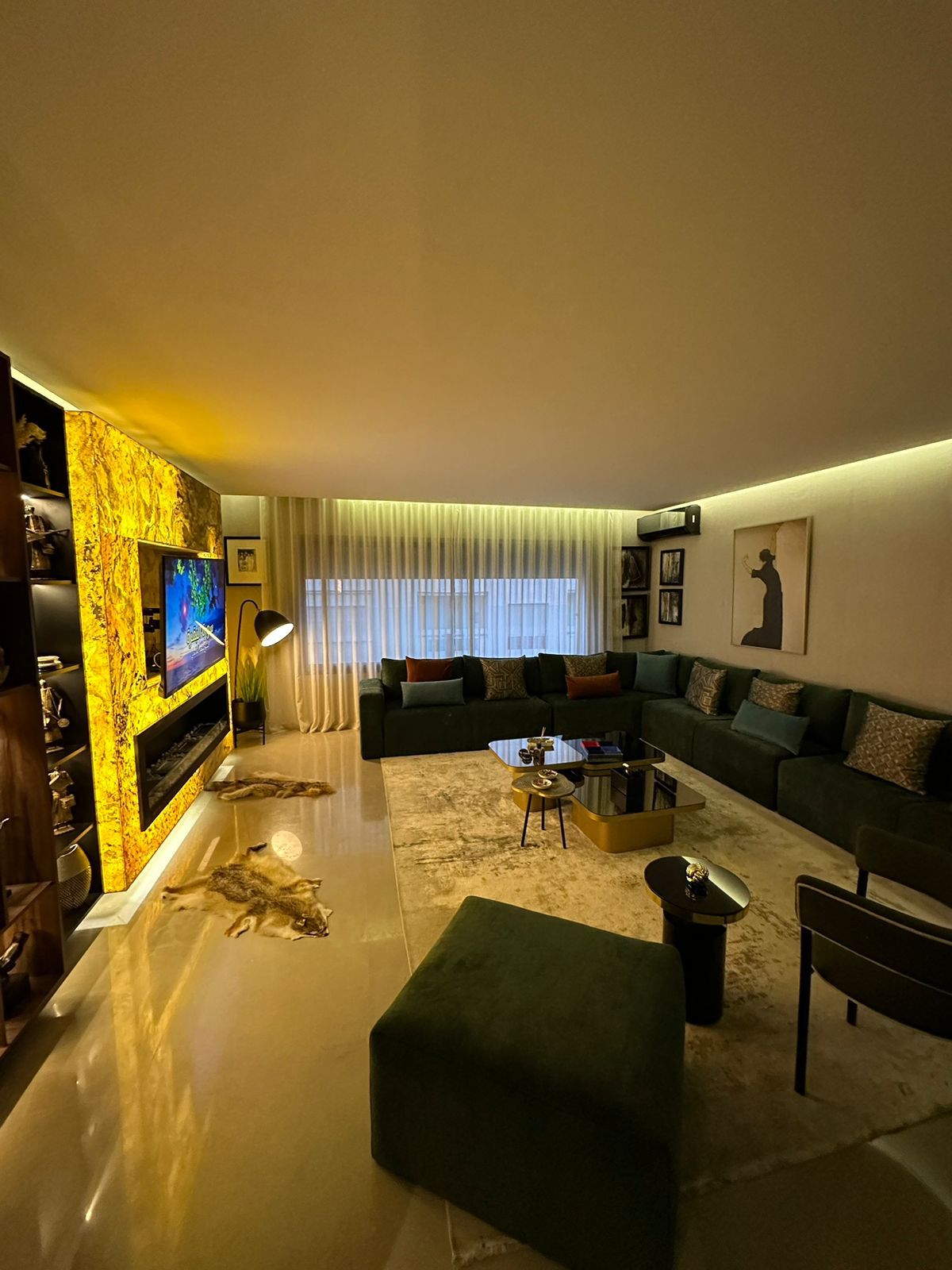 The living area features a spacious green sectional sofa complemented by stylish chairs. A sleek television is mounted above a modern entertainment unit. Natural light filters through sheer curtains, illuminating the glossy floor and the elegant area rug beneath a glass coffee table.