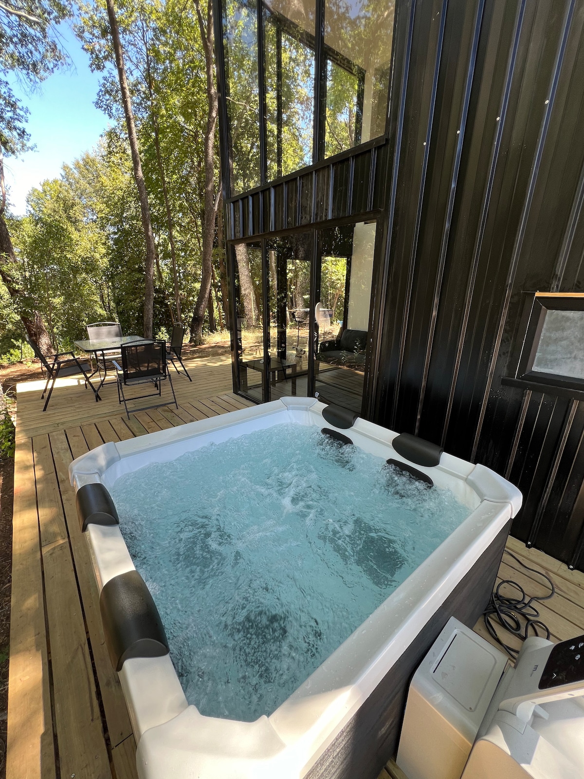 A jacuzzi is situated on a wooden deck, surrounded by lush trees. The structure features large glass walls, providing views of the natural surroundings. Outdoor seating is visible, creating a space for relaxation amid the forest.