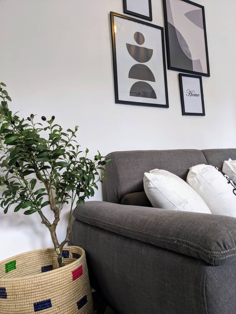 A cozy sitting area is showcased with a gray sofa adorned with white decorative pillows. A small indoor plant is displayed in a woven basket beside the sofa, while art pieces in sleek black frames hang on the wall above.