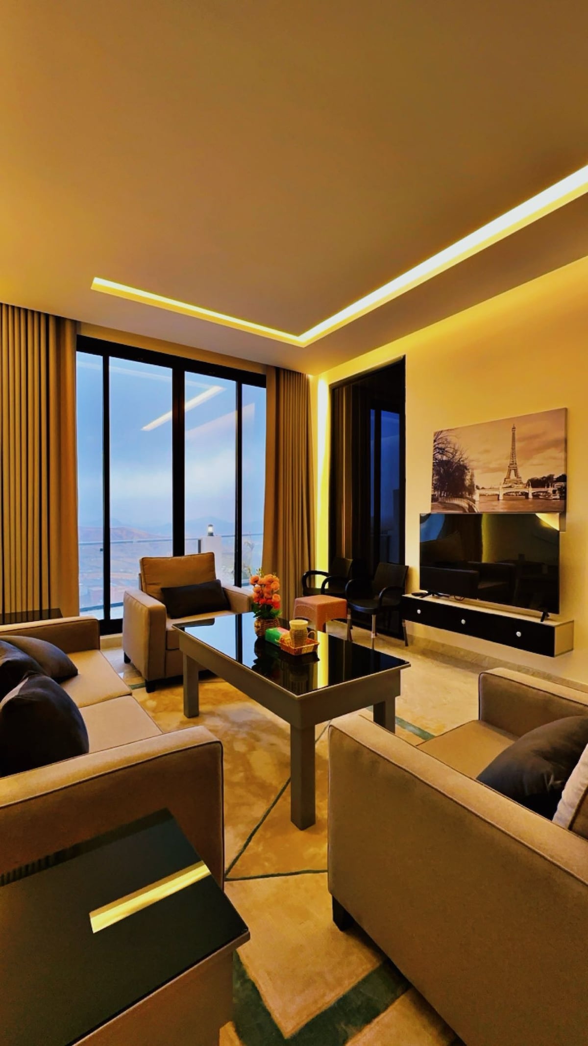 A modern living area is depicted with large glass windows allowing natural light to enter. Comfortable seating is arranged around a sleek coffee table. A flat-screen TV is mounted on the wall, with a decorative photograph adding a touch of elegance.