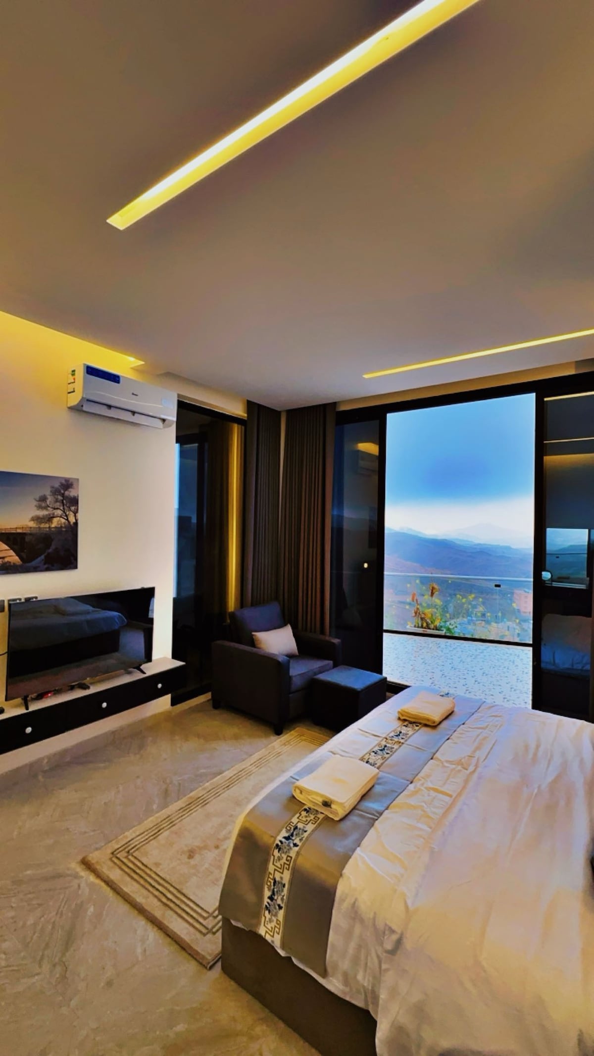 A modern bedroom is equipped with large glass doors that reveal a scenic view of the surroundings. A comfortable chair is positioned near the window, and a sleek television is mounted on the wall. Soft lighting enhances the calming ambiance of the space.