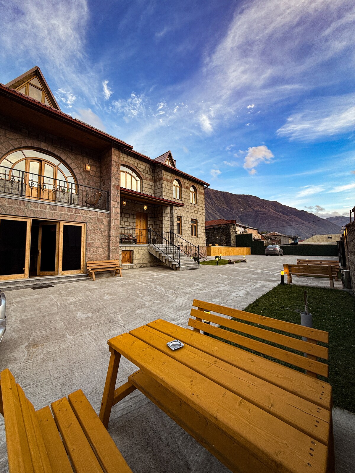 An elegant stone building with large windows is set against a backdrop of mountains. A spacious outdoor area features wooden benches and a well-maintained patio. Soft lighting enhances the inviting ambiance of the exterior space.