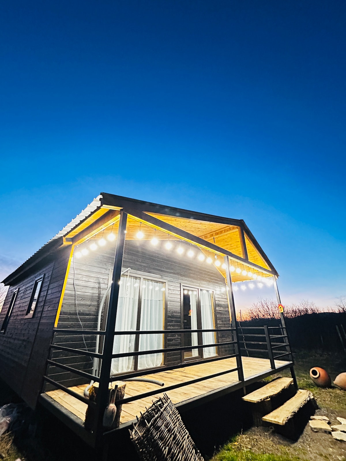 A charming cottage exterior is showcased against a twilight sky, with warm lights adorning the roofline. A spacious porch is visible, featuring railing and wooden decking. Large glass doors lead into the home, enhancing the inviting atmosphere. The surrounding landscape is open and serene.