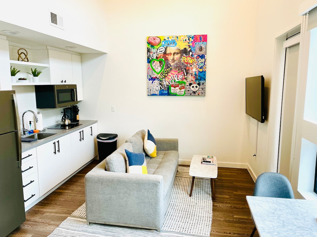 A modern living space features a comfortable gray sofa adorned with colorful throw pillows. A small coffee table sits nearby, and a mounted TV is positioned on the wall opposite. Vibrant artwork adds a pop of color above the sofa, enhancing the sleek kitchen area in the background.