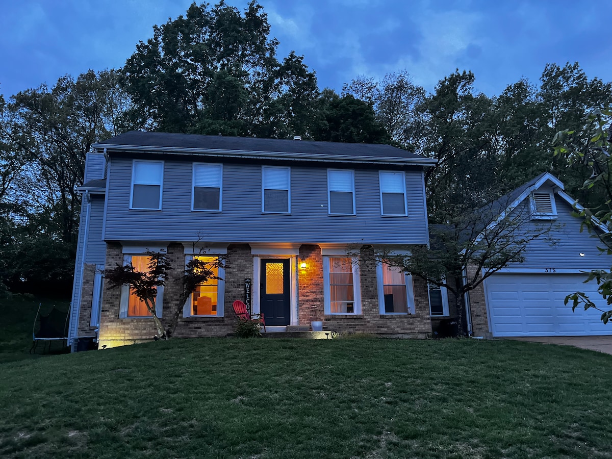 A welcoming two-story family home is presented, featuring a blend of stone and siding. Soft exterior lights illuminate the entryway, which is accented by a red chair. The surrounding grass is neatly maintained, while trees and greenery enhance the natural setting.