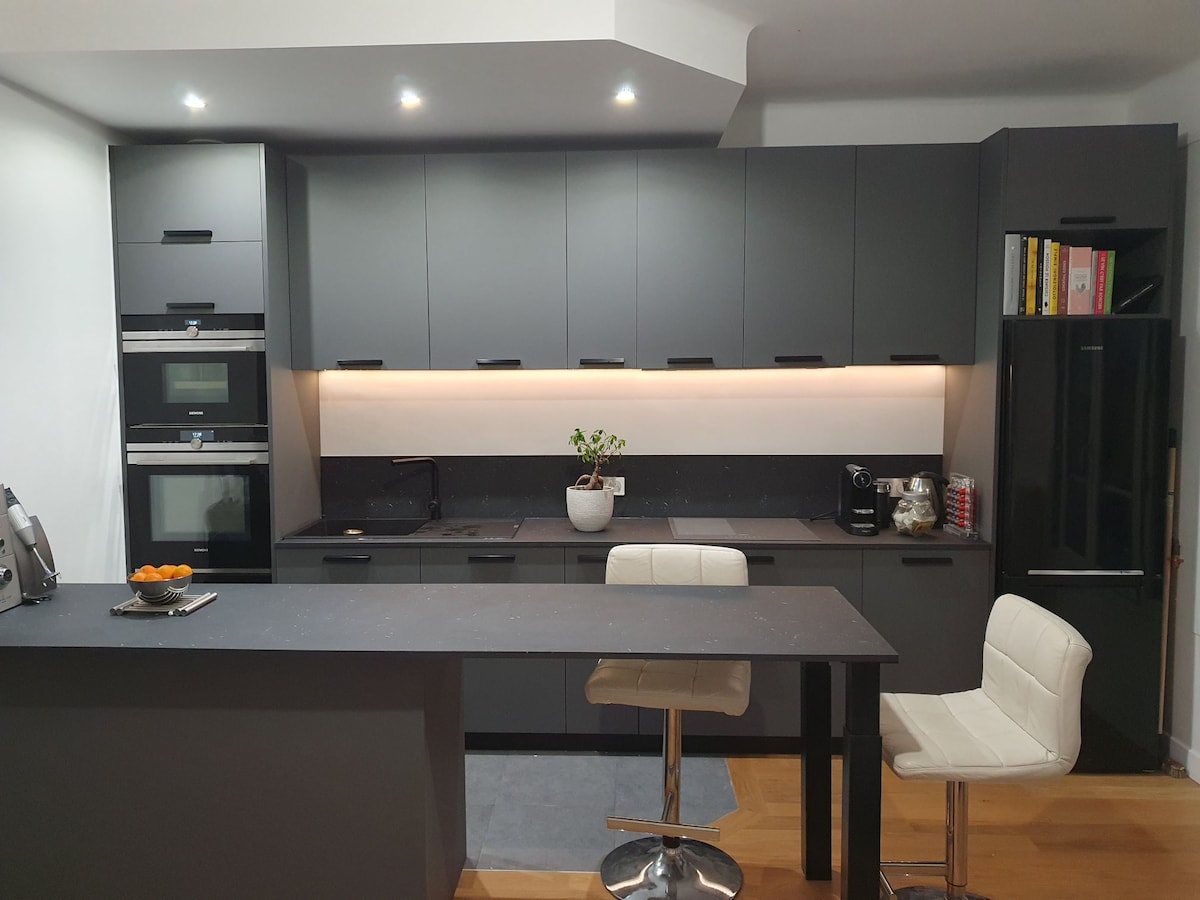 A modern kitchen features sleek grey cabinetry and a smooth countertop. Appliances, including an oven and refrigerator, are neatly integrated. A small bar area with two white stools is present, complemented by warm under-cabinet lighting, creating a functional and welcoming space.