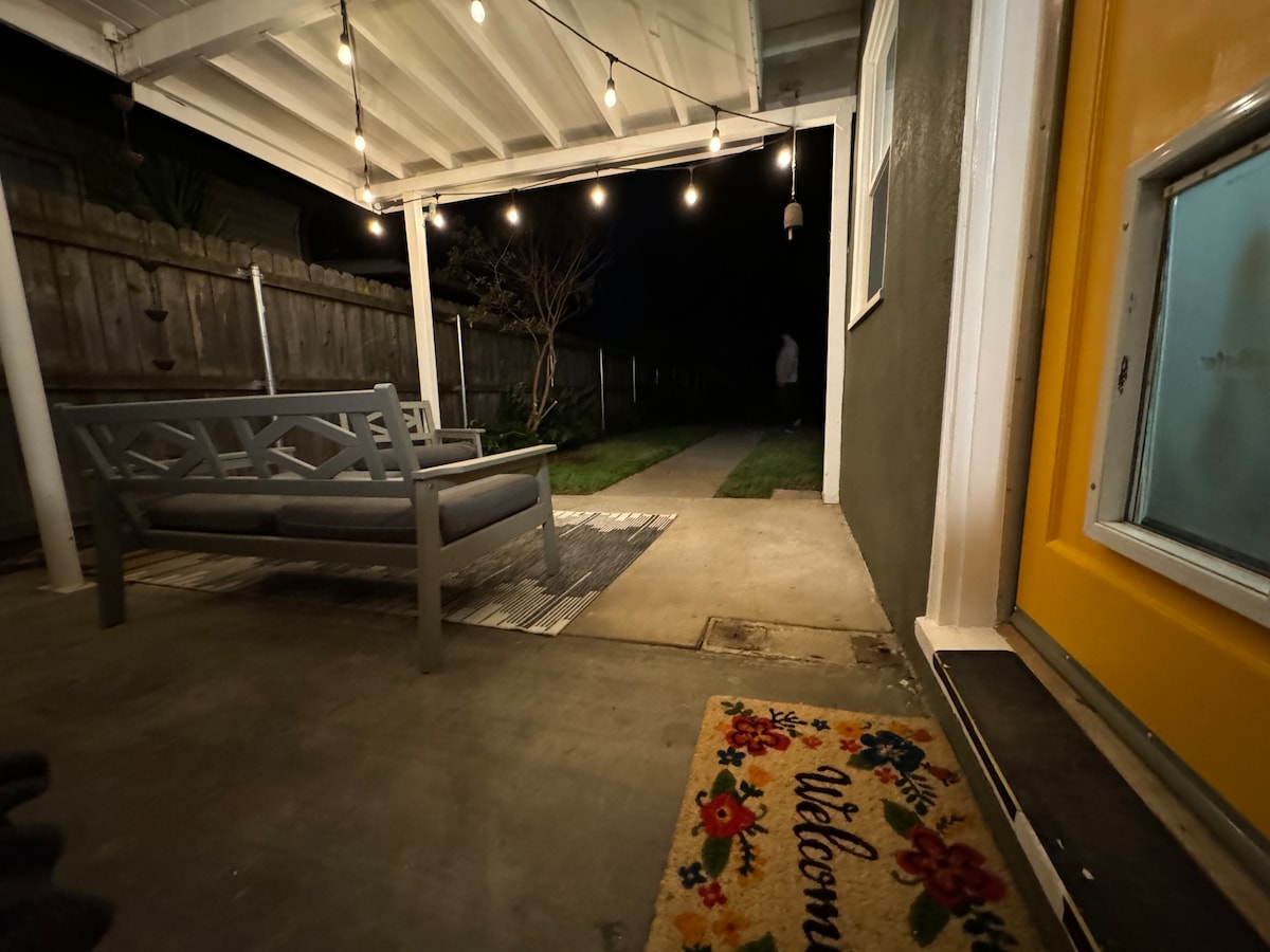 An outdoor seating area is visible, featuring a comfortable gray sofa positioned under string lights. The pathway leads towards a bright yellow door, with a welcome mat placed at the entrance. A backyard space is seen extending in the background.