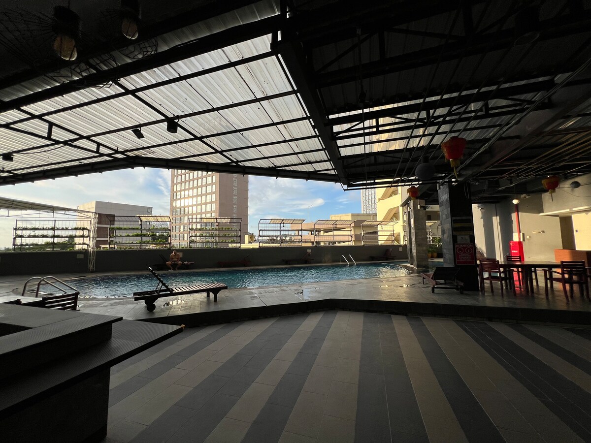 An outdoor pool area is featured under a covered terrace, with a few lounge chairs positioned beside the water. Large windows create an open atmosphere, providing views of surrounding buildings and natural light. A seating area is visible in the background.