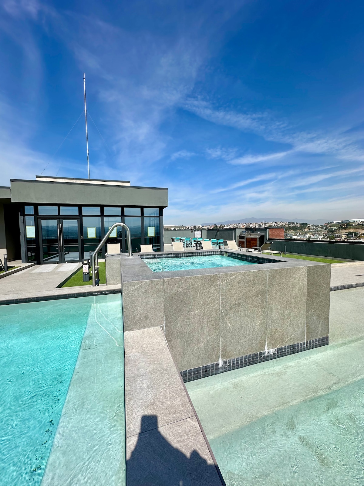 A rooftop pool area is showcased, featuring two connected pools with clear water. Gray stone surrounds and a sleek diving board are visible, alongside modern seating options. Expansive glass walls of the building overlook a scenic view, with blue skies above.