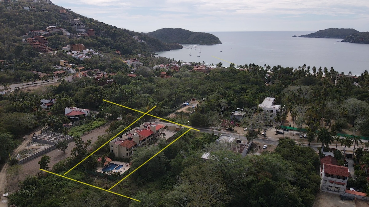 An aerial view of a landscaped area surrounded by greenery, with the ocean visible in the background. A main building with a red-tiled roof is highlighted, along with a swimming pool and several smaller structures. Palm trees and lush vegetation accentuate the surroundings.