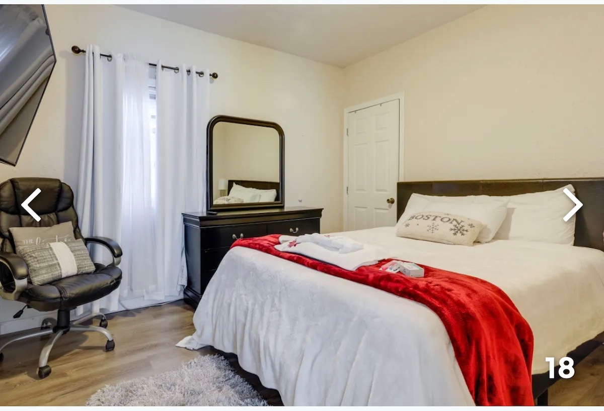 A comfortable bedroom features a spacious bed dressed in white linens and a decorative red throw. A mirrored dresser stands along one wall, accompanied by a task chair for convenience. Natural light filters through the window, which is adorned with white curtains.