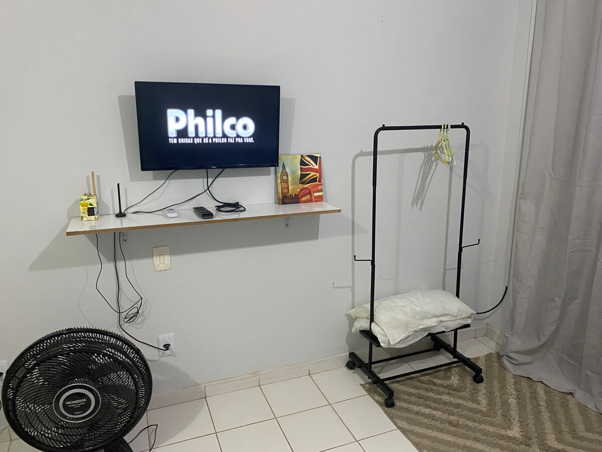 A functional studio space is presented with a mounted television displaying the logo of a brand. A small shelf holds decorative items along with electronic devices. A standing clothing rack is visible, alongside a fan positioned on the tiled floor, contributing to the comfort of the area.