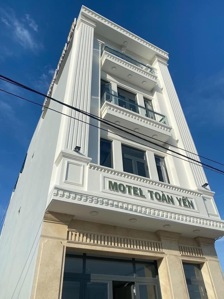 A modern three-story building is framed by a clear blue sky. Prominent columns and decorative moldings enhance the clean, white exterior, while large windows provide ample natural light. The entrance is marked by a glass door, inviting guests into the establishment.
