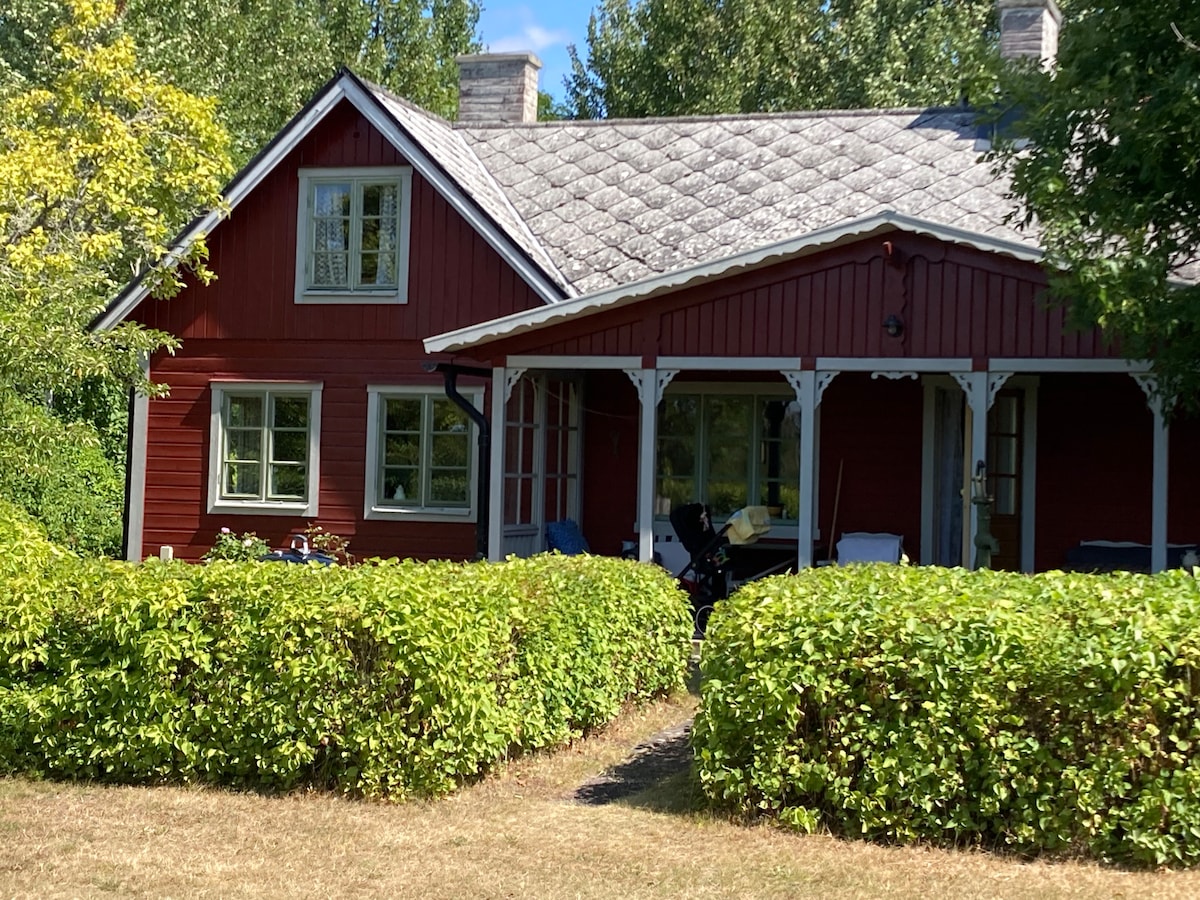 The charming red cottage features a steeply pitched roof and multiple windows, allowing natural light to fill the interior. A covered porch and a pathway surrounded by greenery lead to the entrance, while the tranquil garden offers a blend of outdoor space and privacy.