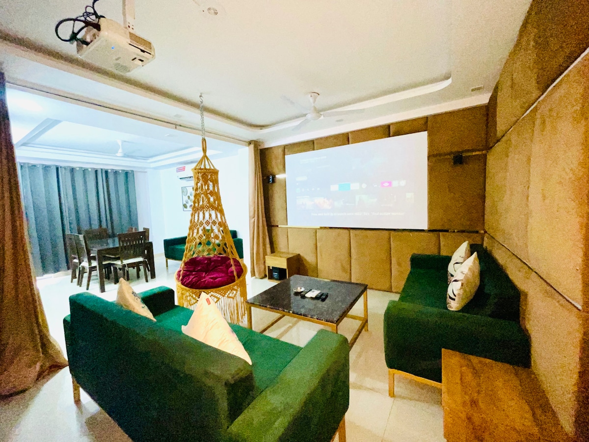 A modern living area is furnished with deep green sofas arranged around a central table. A projector screen covers one wall, while a decorative hanging chair adds charm. Natural light filters through sliding doors leading to an outdoor dining area, enhancing the space's openness.