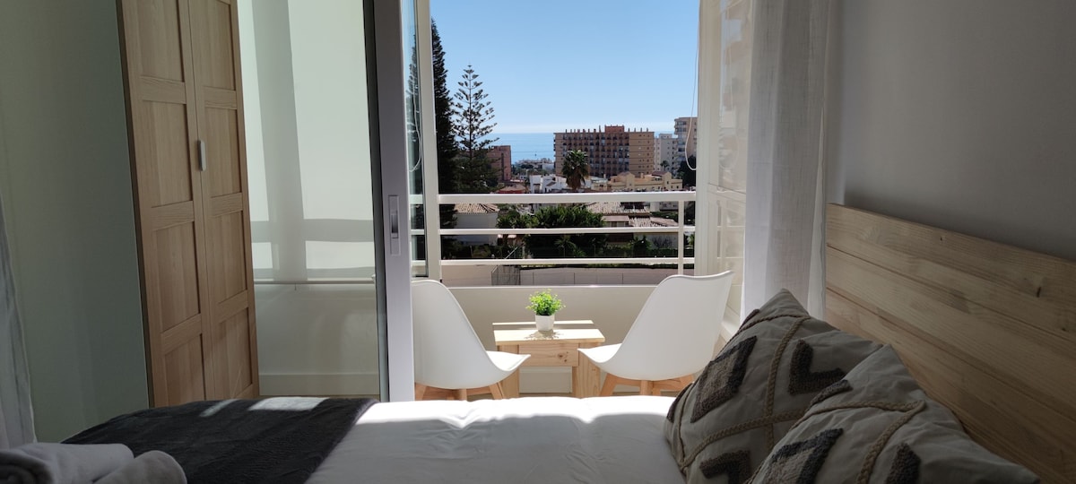 A bright bedroom features a spacious bed and large sliding doors that open to a small balcony. Two minimalist chairs and a small table provide a relaxing outdoor space, with views of the surrounding area and the sea in the distance.
