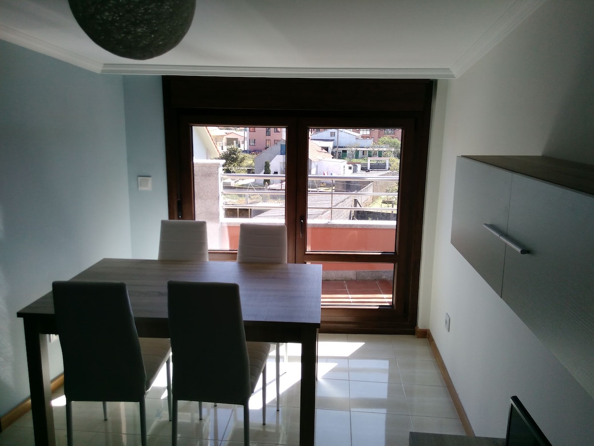 A dining area with a wooden table and four white chairs is illuminated by natural light from large windows. The view outside includes rooftops and greenery, framed by the warm wood of the window. Light tiles reflect the brightness within the room.