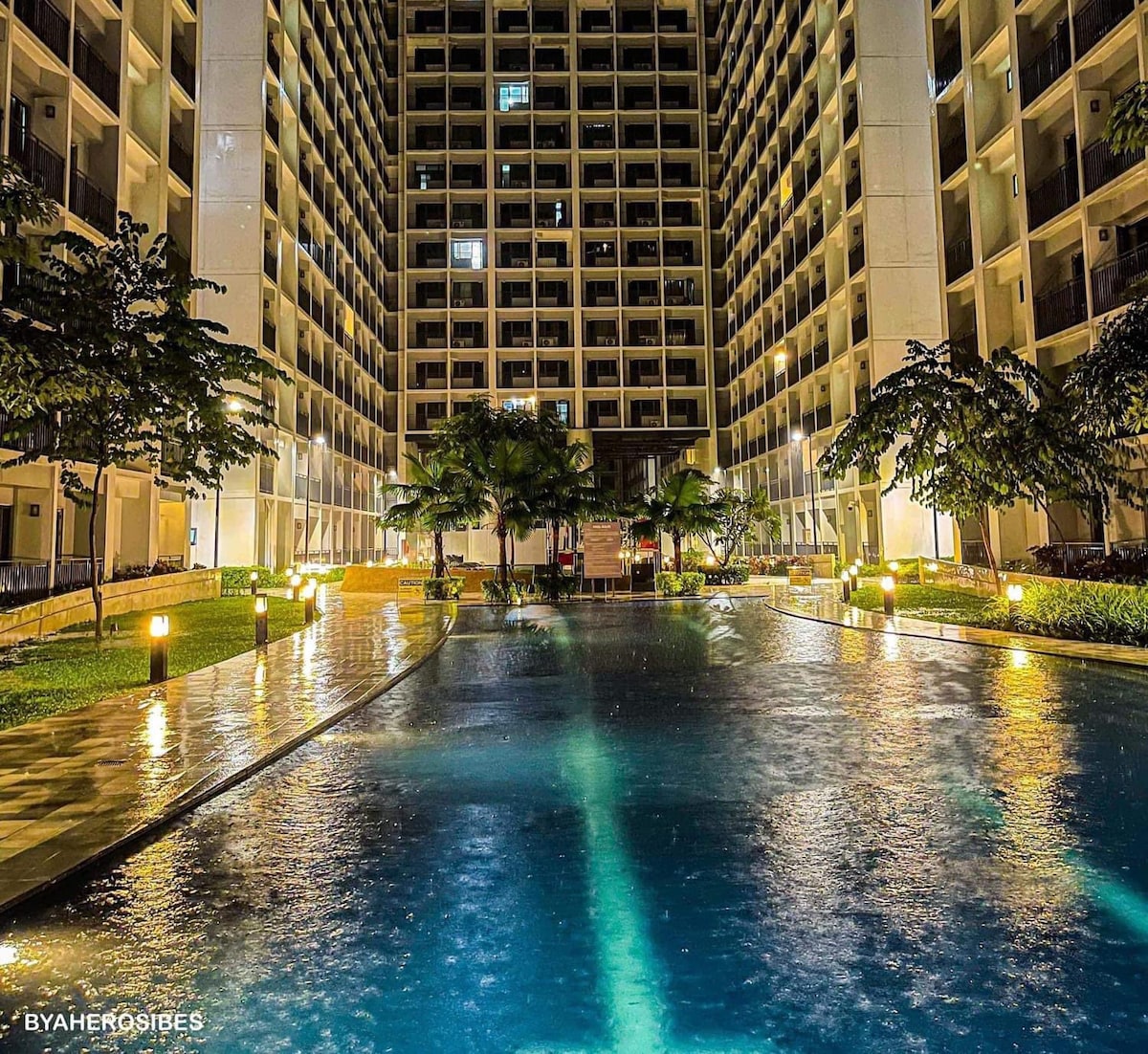 An inviting swimming pool is surrounded by a well-maintained courtyard. Soft lighting reflects off the water, creating a serene atmosphere. Towering buildings frame the pool area, and palm trees are positioned strategically throughout the landscaped space, enhancing the overall tropical ambiance.