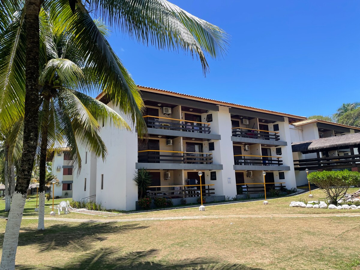 A three-story building is surrounded by lush palm trees and neatly trimmed grass. Balconies are visible on the upper floors, and air conditioning units are mounted on the exterior walls. Clear blue skies enhance the serene environment of the property.