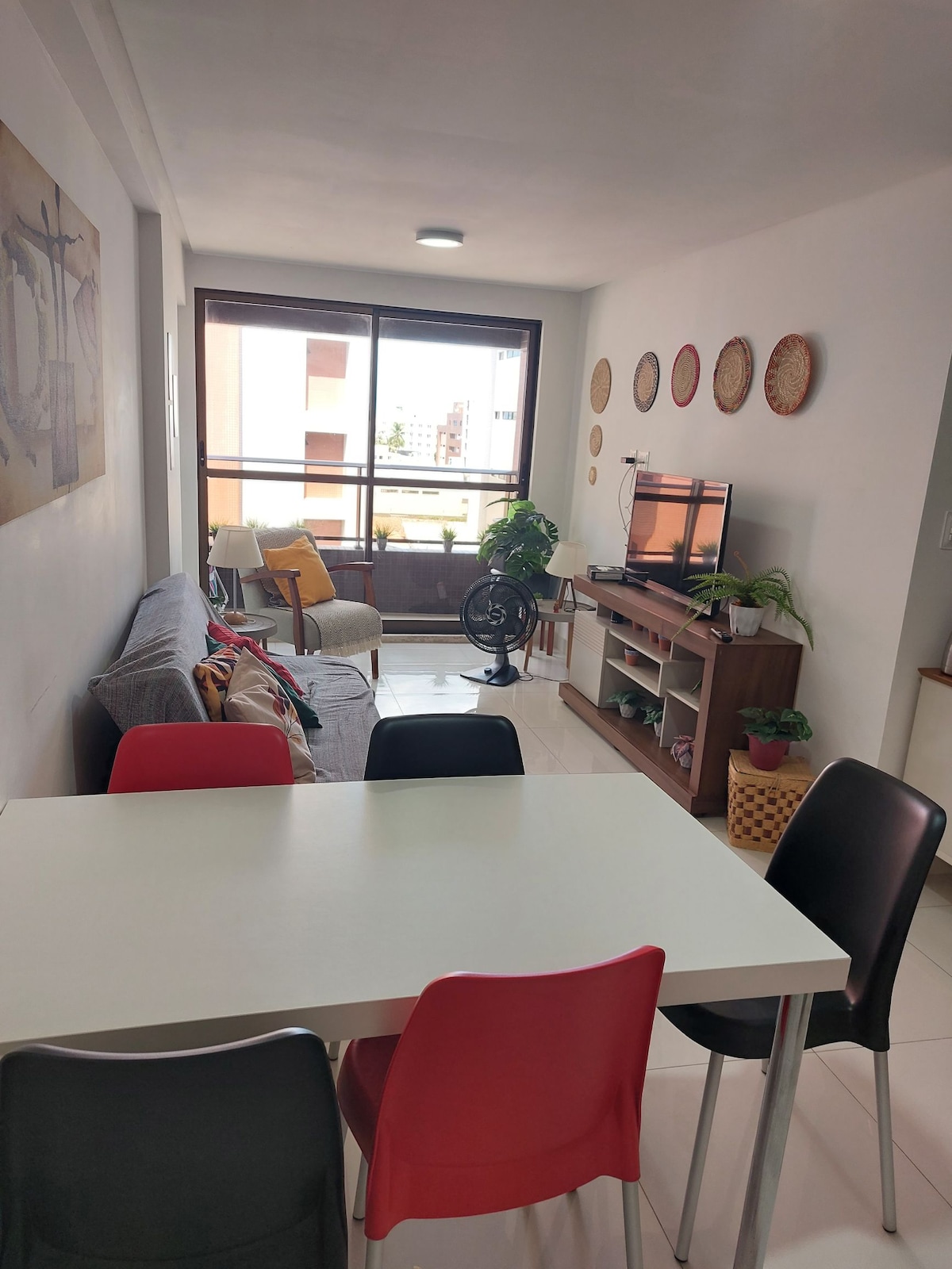 A bright living area features a comfortable sofa and a television unit. A small dining table with colorful chairs adds a lively touch. Natural light flows in through the large window, enhancing the spacious feel. Decorative wall hangings bring subtle texture to the space.