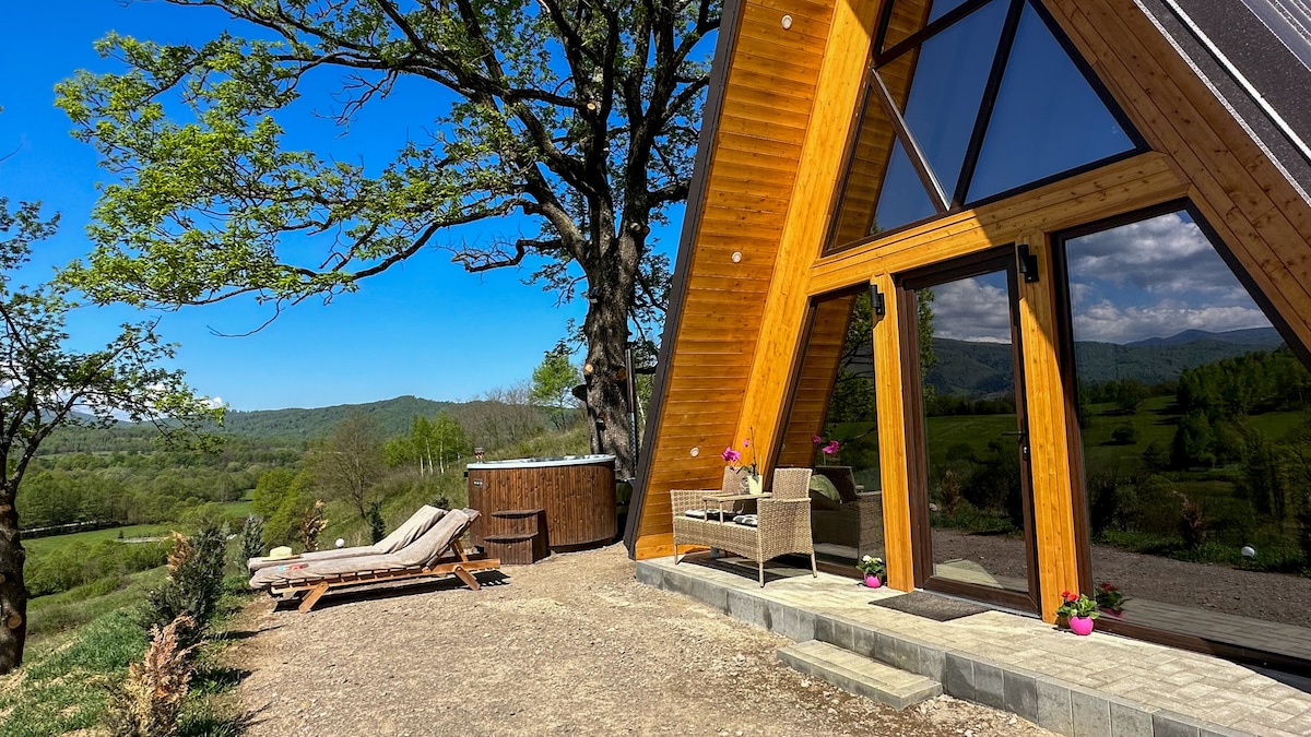 An A-frame structure features large glass doors, allowing natural light to fill the interior. A relaxing seating area with loungers sits nearby, overlooking a scenic view. A hot tub is positioned on the side, surrounded by natural landscaping and set against a backdrop of rolling hills.