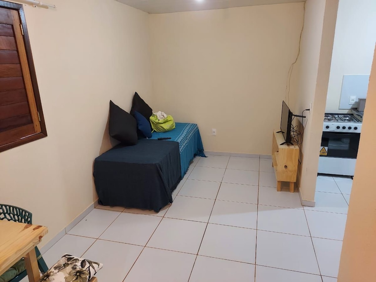 A simple living area features a single bed with dark bedding and two large cushions in contrasting colors. A small table and chair are present nearby. A television is positioned on a wooden stand opposite the bed, with a kitchenette visible in the background.