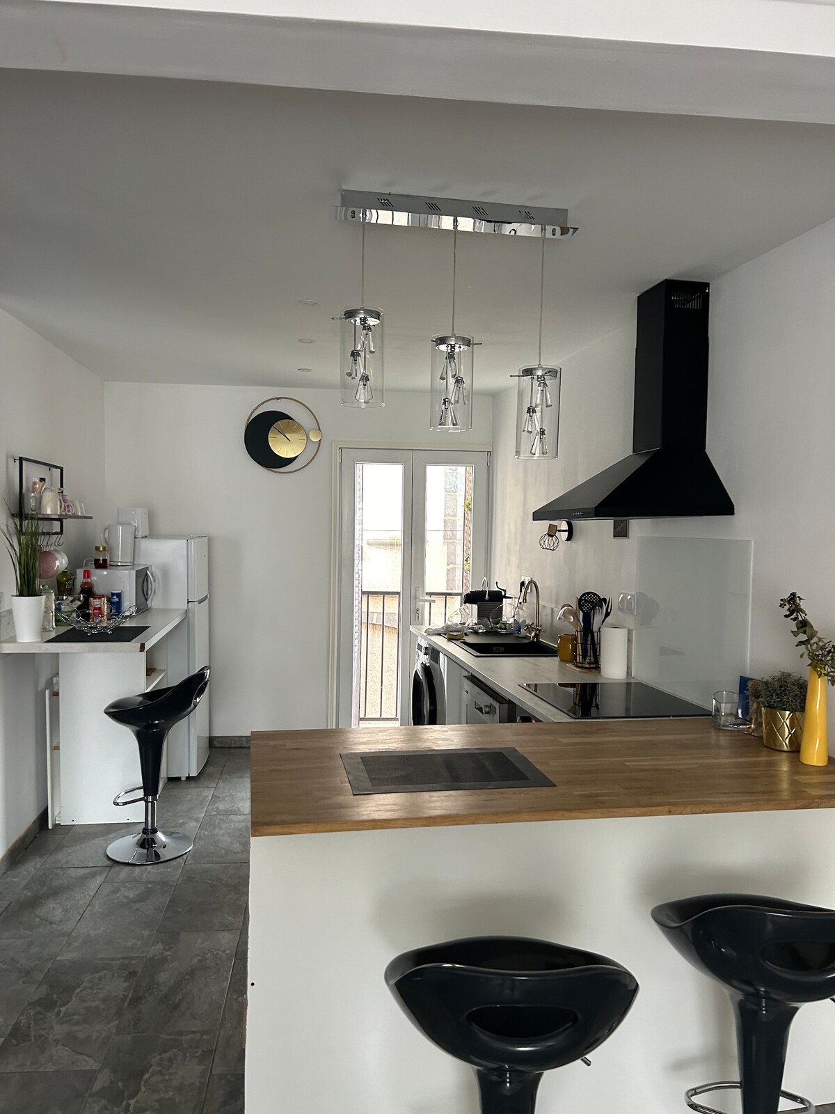A modern kitchen area features a sleek countertop with two black bar stools. Stainless steel appliances, including a refrigerator and oven, are visible. Large glass doors lead to a balcony, allowing natural light to fill the space. Simple decor adds a contemporary touch.